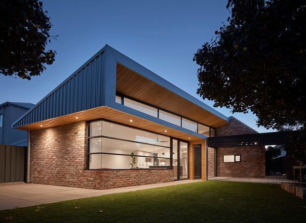 Twilight exterior view of a contemporary two-storey residence with a strong angular roof form, exposed red brick walls, blue-grey vertical metal cladding, and a warm timber-lined soffit. Large glazed openings reveal a bright interior, while recessed downlights illuminate the covered entry and lawn. A dark roofed carport or connecting canopy is visible to the right, framed by mature trees.