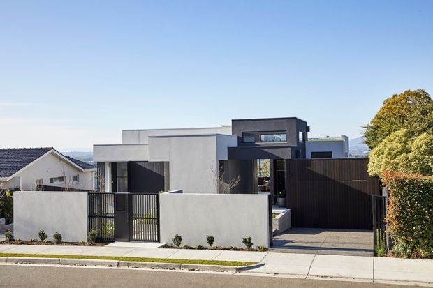 Street-facing view of a modern two-storey residence with a flat-roofed geometric form, smooth light rendered walls, dark vertical screening and garage/entry elements, a small enclosed front garden, and a concrete driveway. The home sits in a suburban setting with neighbouring houses visible.