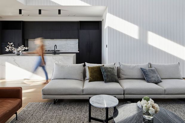 Bright contemporary open-plan living area with a large light-grey sectional sofa in the foreground, paired round coffee tables and a textured rug. Behind it is a compact kitchen with dark timber cabinetry, a white marble-look island and splashback, and a black tap. A white vertical-ribbed feature wall and strong sunlight create a crisp, refined interior.