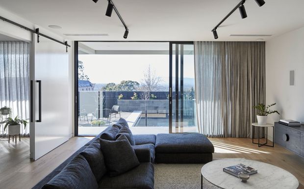 Contemporary living room with a large dark sectional sofa, round coffee table, timber flooring, full-height glazing and curtains opening onto a balcony or terrace with an outdoor pool area beyond. Black track lighting, minimalist cabinetry and neutral finishes create a clean, refined interior.