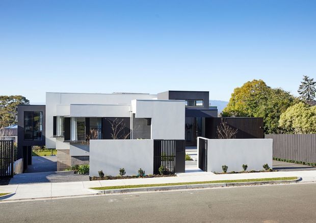 Street-level exterior view of a contemporary multi-volume residence with crisp white rendered walls, dark charcoal feature volumes, extensive glazing, vertical black screening, and a low front boundary wall. The house sits along a paved suburban street with landscaped planting and mature trees beyond.