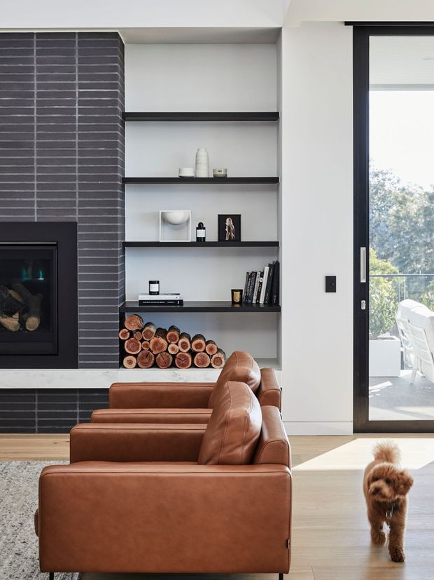 A bright contemporary living room featuring a dark horizontal-brick fireplace, built-in open shelving with decorative objects, books and stacked firewood, and two cognac leather lounge chairs. A large black-framed glazed door opens to an outdoor terrace, while pale timber flooring and white walls create a clean, warm interior.