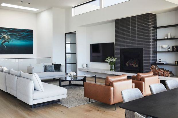Bright contemporary living room with a high ceiling and clerestory windows, furnished with a large light-grey sectional sofa, cognac leather armchairs, a dark dining table, and pale upholstered dining chairs. A black tiled fireplace wall with built-in TV and open shelving forms the main feature, with timber flooring, a large rug, and coastal-themed artwork.