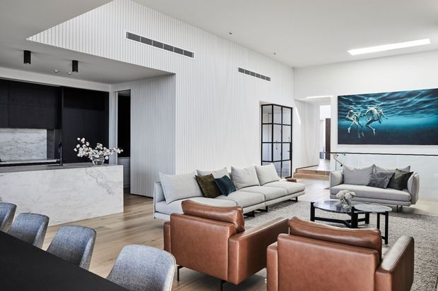A bright, open-plan contemporary living and dining area with a large light sectional sofa, brown leather armchairs, a dark dining table with upholstered chairs, and a marble-look kitchen island. White vertically ribbed wall and ceiling panels, black-framed glazed doors, pale timber flooring, and a large ocean-themed artwork create a refined monochromatic interior.