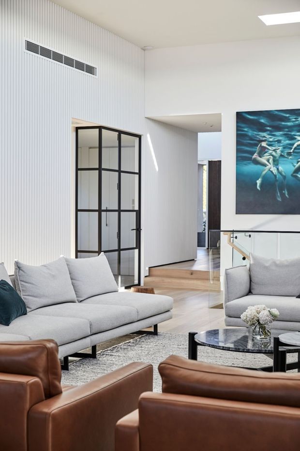 Bright contemporary living room with light grey sofas, brown leather armchairs, a black glass coffee table, and a grey rug. The room features white vertical fluted wall panelling, light timber flooring, black-framed glazed internal doors, a glass stair balustrade, and a large underwater artwork. A hallway and upper-level opening are visible beyond the seating area.