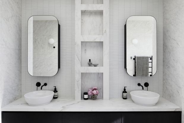 Symmetrical contemporary bathroom vanity with two white vessel basins, black tapware, rounded mirrored medicine cabinets, white marble-effect wall and benchtop surfaces, recessed display niches, and dark lower cabinetry. The space has a clean, refined monochromatic scheme with subtle warm floral styling.