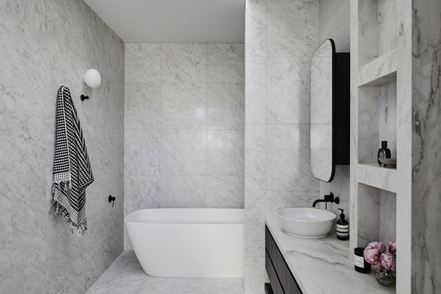 Contemporary bathroom clad in white-grey marble-look surfaces, featuring a freestanding white bathtub, vessel sink on a marble vanity, black fixtures, a rounded rectangular mirror, recessed shelving, and monochrome textiles with small floral accents.