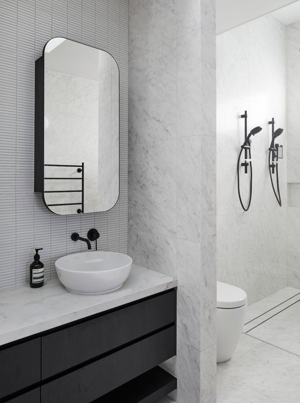 Contemporary monochrome bathroom featuring a floating dark vanity with a white vessel basin, black wall-mounted tapware, a rounded-arch mirror, pale marble-look wall and floor tiles, and a glass-enclosed shower with black fixtures. A toilet is visible beyond the partial dividing wall.