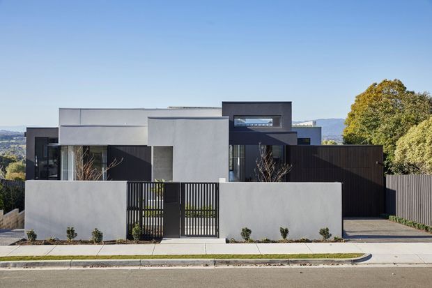 Street-facing view of a contemporary multi-level residence with strong geometric forms, flat roofs, dark vertical cladding, smooth light rendered walls, and large glazed openings. A low rendered boundary wall and black metal pedestrian gate define the front, with a paved driveway and sparse landscaping. Distant hills and mature trees are visible beyond the property.
