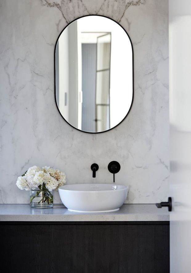 A refined bathroom vanity with a white vessel basin set on a dark timber-look cabinet, black wall-mounted tapware, and a rounded black-framed mirror. The wall is clad in pale marble with subtle grey veining, creating a high-contrast, luxurious monochrome composition. A small vase of white flowers sits beside the basin.