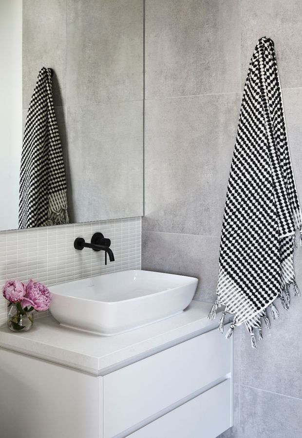 A contemporary bathroom vanity with a white vessel basin set on a pale countertop and white built-in cabinetry. Large light-grey concrete-look wall tiles are paired with a narrow horizontal white mosaic-tile backsplash. A matte black wall-mounted tap and black-and-white patterned towels provide contrast, while a small vase of pink flowers sits beside the basin.