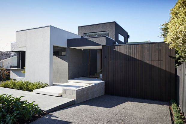 Street-facing view of a contemporary multi-level residence with clean geometric volumes, white and dark charcoal rendered walls, a projecting flat-roof entry canopy, dark vertical timber fencing, and a paved driveway. Landscaping is minimal and the facade features broad horizontal and clerestory-style glazing.