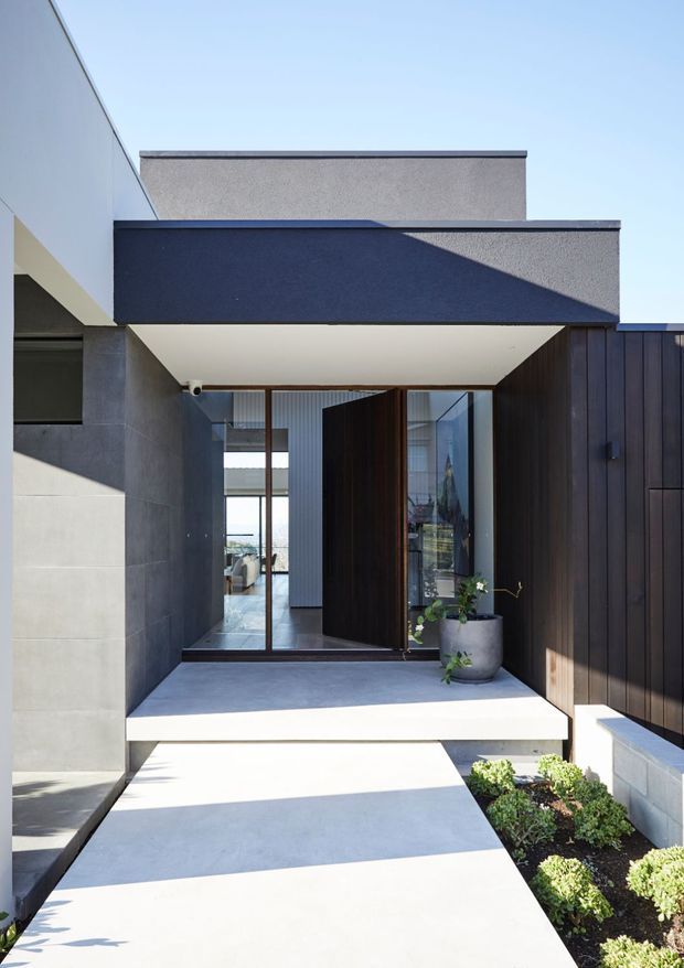 Contemporary residential entrance viewed from an exterior walkway, featuring crisp geometric volumes, a deep dark projecting canopy, pale rendered and concrete surfaces, dark vertical timber cladding, large glazed entry doors, and minimalist landscaping with clipped shrubs and a potted plant.