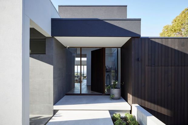 Contemporary flat-roofed residential entrance with geometric rendered walls, dark vertical timber screening, a recessed glazed doorway, and a concrete-paved path leading to the entry. Strong sunlight creates crisp shadows across the minimalist facade.