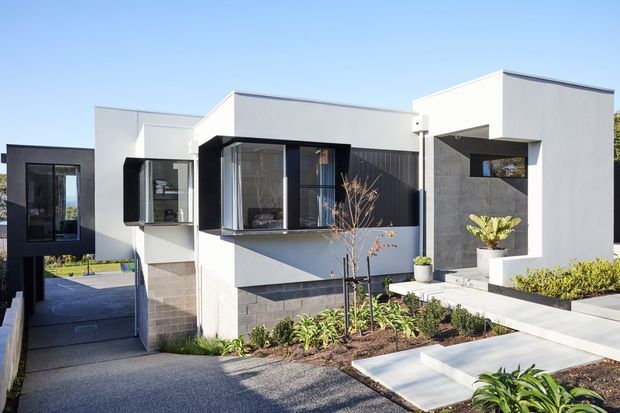 Contemporary single-storey residence with a strongly geometric, cubic composition. The facade combines white rendered wall planes, dark charcoal cladding and window frames, large glazed openings, and a paved landscaped front approach with raised garden beds and concrete steps.