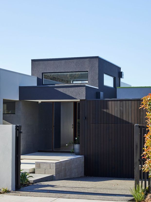 A modern multi-level residential facade viewed from the driveway. The house features dark textured rendered walls, crisp rectilinear massing, large horizontal windows, a projecting entry canopy, dark vertical timber fencing and a paved front approach. A small garden planting edge and bright sky are visible around the building.