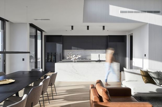 A bright, open-plan contemporary kitchen, dining and living area. The space features a large white stone kitchen island with integrated sink, dark built-in cabinetry, expansive glazing, a black dining table with upholstered chairs, and a white sofa with leather seating. Strong sunlight creates geometric shadows across the light timber floor and vertically ribbed wall surfaces.