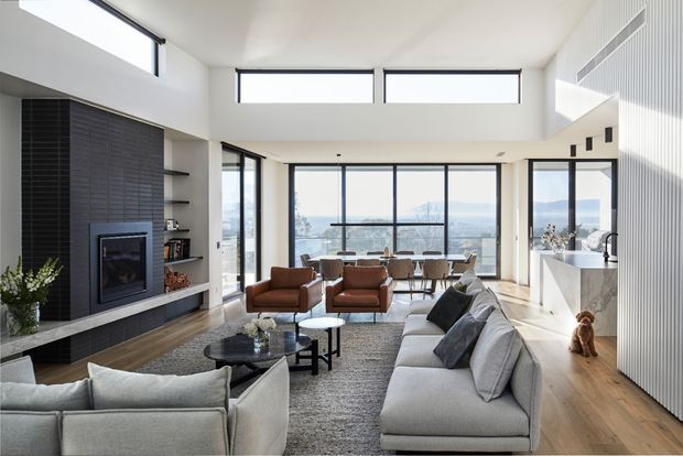 Bright, spacious open-plan living room with a high ceiling and expansive floor-to-ceiling glazing overlooking a distant landscape. The room features a large grey sectional sofa, additional lounge chairs, a dining area, warm timber flooring, a dark tiled fireplace wall, and a white vertically ribbed feature wall with integrated shelving and joinery.