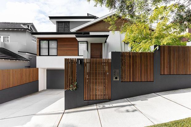 Street-facing view of a contemporary multi-storey residence with a pitched gable roof, white rendered walls, warm timber cladding and vertical timber privacy fencing. The property includes a dark grey garage door, gated entry, driveway and a partially visible neighbouring dwelling.