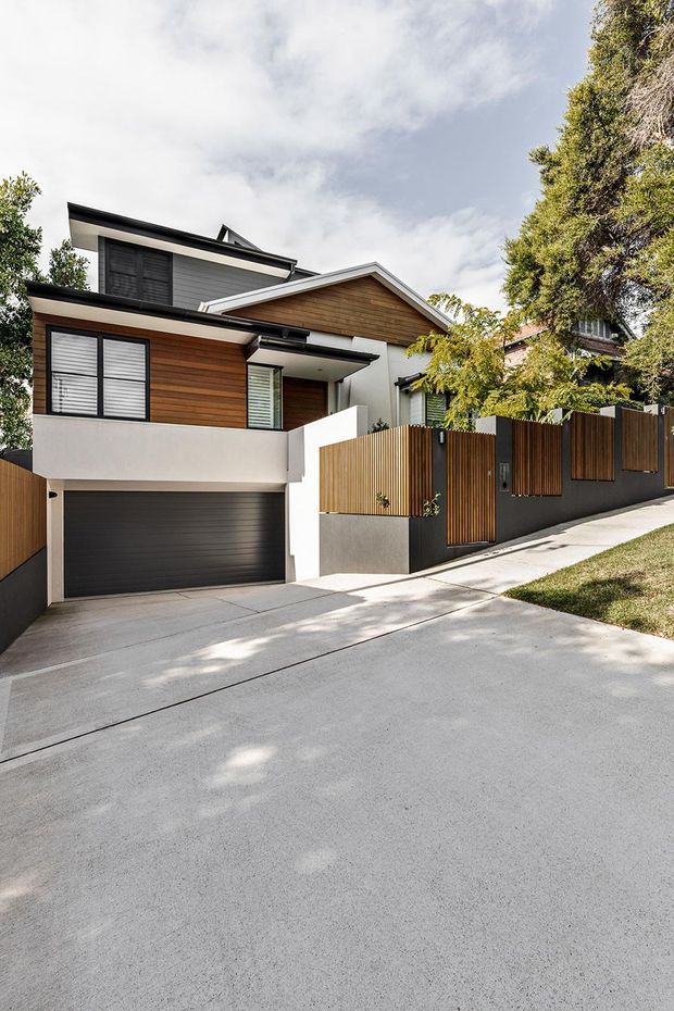 Street-facing exterior of a contemporary multi-level residence on a sloping site, featuring horizontal timber cladding, white rendered walls, dark garage door and window frames, pitched roof forms, and a timber privacy fence above a broad concrete driveway.