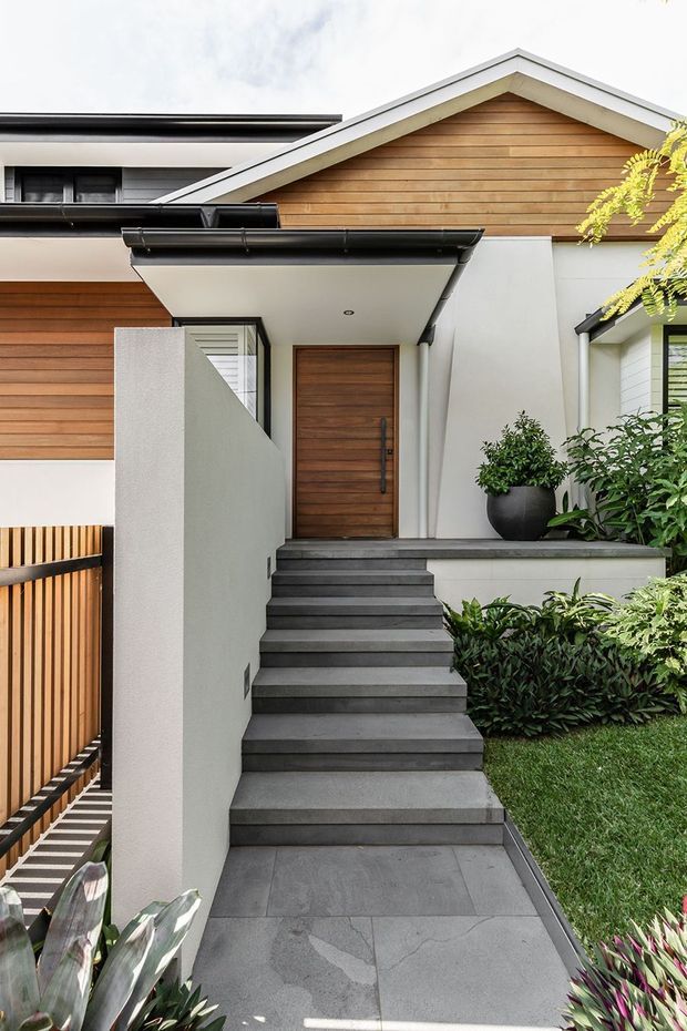 A contemporary residential front entry viewed from the garden, with a broad dark tiled stairway leading to a timber front door beneath a covered porch. The facade combines white rendered walls, warm horizontal timber cladding, black gutters and trim, and a pitched gable roof. Lush planting, a clipped hedge, and a timber slat fence frame the approach.