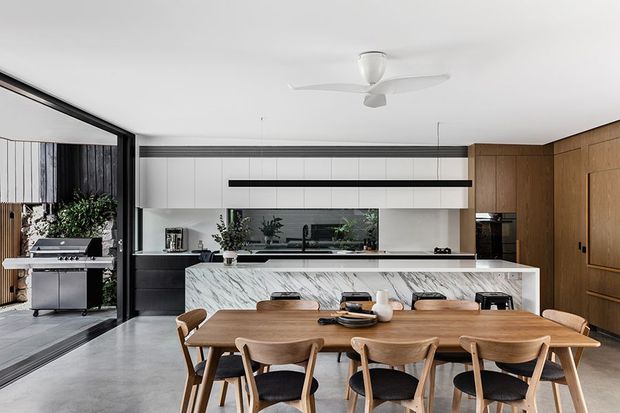 A spacious contemporary open-plan kitchen and dining area with a long marble-look island, white and timber cabinetry, integrated appliances, and a large wooden dining table with matching chairs. Full-height glazing opens toward an outdoor barbecue area, while a white ceiling fan and linear pendant-style lighting contribute to the clean modern interior.