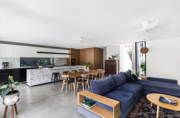 Open-plan contemporary living, dining and kitchen area with a large marble-look island, white integrated cabinetry, warm timber joinery, concrete-look flooring, timber dining furniture and a deep navy sectional sofa. The bright room includes extensive glazing, indoor plants, ceiling fans and a patterned rug.