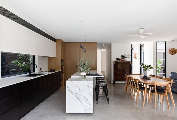 Open-plan contemporary kitchen and dining area with a long black and white kitchen run, marble-look island, timber wall cabinetry and paneling, integrated appliances, and bar seating. A timber dining table and chairs sit adjacent to the kitchen, with a living area visible beyond. The space has polished concrete flooring, a white ceiling, abundant natural light, and restrained modern detailing.