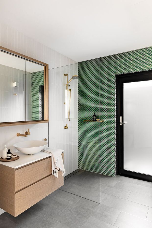 Contemporary bathroom featuring a floating light-wood vanity with a white vessel basin, large timber-framed mirror, brushed brass tapware and shower fittings, white vertically tiled walls, and a striking green small-format tiled feature wall. A black-framed glazed door and large grey floor tiles complete the refined, high-contrast scheme.