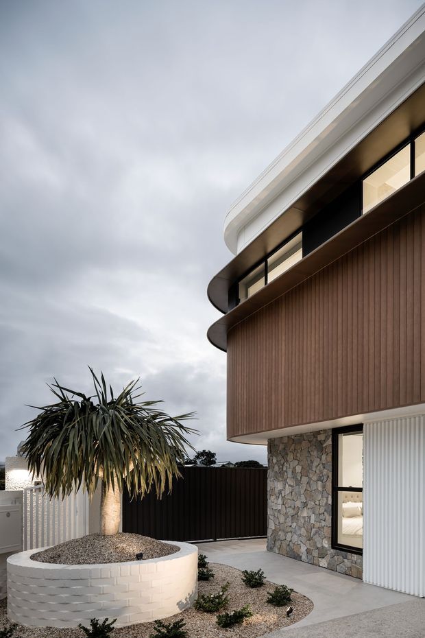 Exterior architectural photograph of a contemporary multi-level residence, focusing on a rounded projecting upper storey with dark-framed glazing, vertical timber-look cladding, natural irregular stonework, and smooth white roof eaves. A landscaped courtyard includes a circular white brick planter with a sculptural tree, gravel beds, low planting, concrete paving, and dark fencing. The image is captured under an overcast sky.