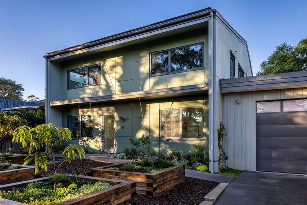 Contemporary two-storey home with a compact, rectilinear form, dark metal roof, vertical grey cladding, large windows, and an integrated single garage. The landscaped front garden uses raised timber-edged planter beds and dark mulch, with a concrete driveway and pathway leading to the entry.