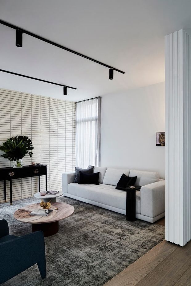 A refined contemporary living room with a white sectional sofa, dark accent cushions, black console table and side tables, a round stone or marble coffee table, and a large textured grey rug. White walls are paired with a full-height linear feature wall, sheer curtains, timber flooring, and black linear track lighting. Vertical white fluted panelling forms a prominent architectural screen at the right edge.