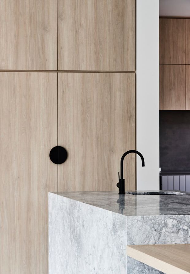 Close-up architectural detail of a contemporary kitchen featuring full-height light timber cabinetry with flush fronts and a round black pull, a substantial pale veined stone island with waterfall edge, and a matte black gooseneck kitchen tap. Dark timber cabinetry and a dark splashback are visible in the background.
