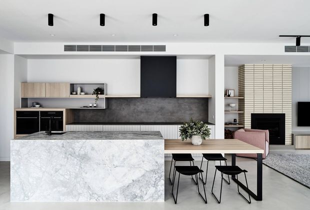 Contemporary open-plan kitchen and dining area with a large white marble-look island, light timber cabinetry, dark stone backsplash, integrated black appliances, and a timber dining table with black chairs. A fireplace and lounge area are visible beyond the dining zone, with recessed linear lighting and minimalist detailing throughout.