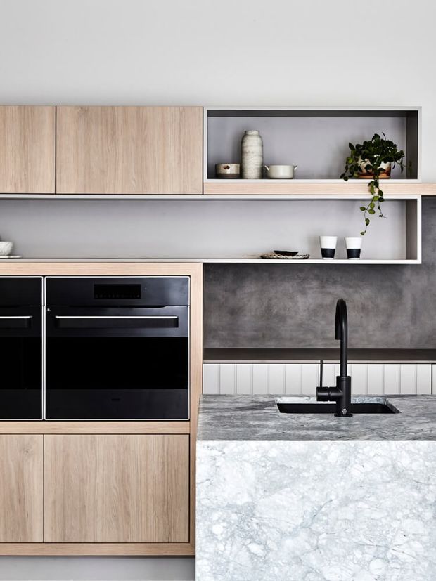 Contemporary kitchen detail featuring light timber cabinetry, integrated black appliances, open display shelving, a dark textured splashback, and a substantial pale veined stone island benchtop with a matte black sink and tap. Minimal ceramic decor and trailing greenery add a restrained natural accent.