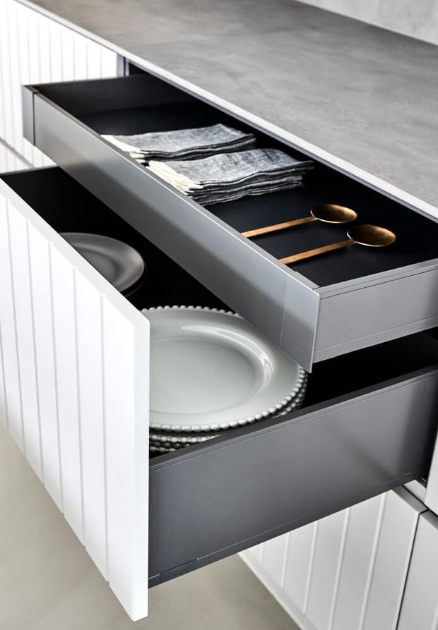 Close-up of open built-in drawers in a white fluted cabinet, showing organised tableware, folded napkins and gold-toned spoons. The drawers have dark interiors and sit beneath a grey stone-look countertop, highlighting kitchen or dining storage joinery.