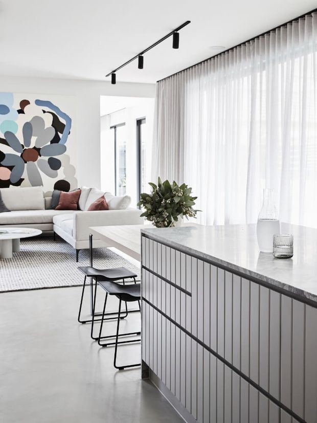 A bright contemporary open-plan living and kitchen space. A white tiled island with dark stone benchtop and black-framed stools dominates the foreground, while a white sectional sofa, textured rug, sheer full-height curtains, abstract artwork, and indoor greenery occupy the living area beyond. A slim black linear track light runs across the ceiling, and the palette is predominantly white, light grey, and black with muted blush and blue accents.