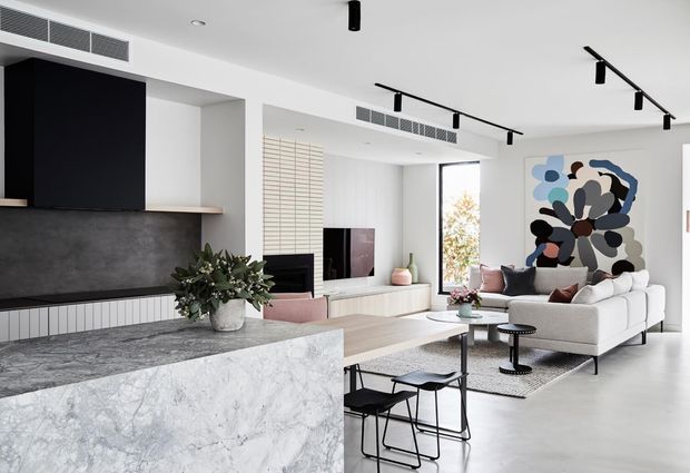 A bright, open-plan contemporary living area adjoining a kitchen. The room features a large marble-look island, dark textured fireplace or media wall with linear built-in joinery, pale timber flooring, white walls, track lighting, a light sectional sofa, black-framed occasional furniture, and a large abstract artwork.