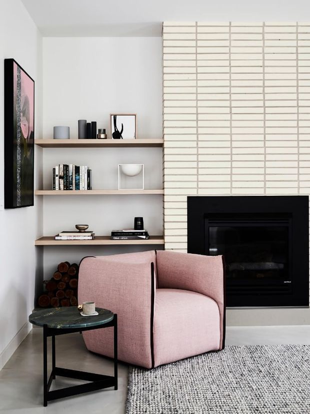 A refined living room corner with a blush-pink upholstered lounge chair, black-framed round side table, woven grey rug, floating timber shelves displaying books and decor, stacked firewood, and a black fireplace set into a white linear brick feature wall. The palette is light and neutral with soft pink upholstery and strong black accents.