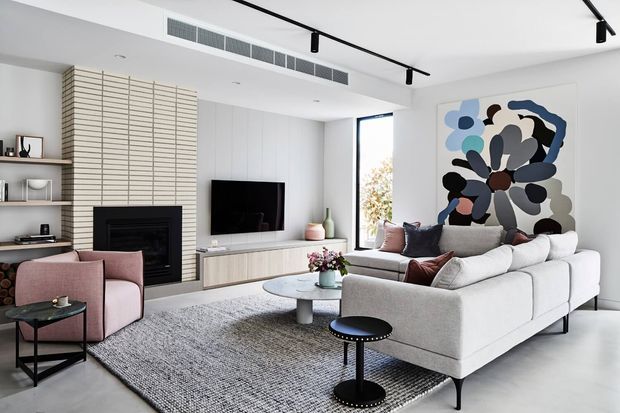 Bright contemporary living room with a large light sectional sofa, blush accent chairs, black metal coffee and side tables, a textured grey area rug, pale built-in media cabinetry, and a linear black track light. A black fireplace is framed by a pale horizontal slatted feature wall, with a large abstract artwork adding colour to the neutral scheme.
