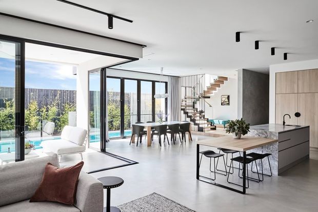 Bright open-plan contemporary living, dining and kitchen area with floor-to-ceiling black-framed glazing opening to a pool terrace. The space features a pale neutral palette, polished concrete-style flooring, a stone kitchen island with timber dining extension, minimalist black track lighting, and an open floating staircase with slim metal balustrades. A partial living area and outdoor pool are visible through the glazing.