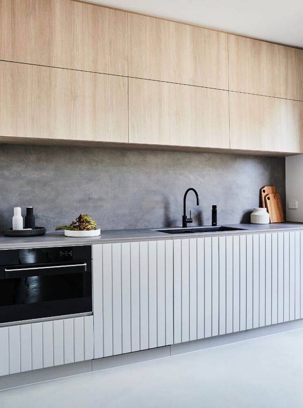 A contemporary minimalist kitchen featuring light timber overhead cabinetry, vertically fluted pale cabinetry below, a dark grey stone-look benchtop and matching textured splashback, a black integrated oven, and a black gooseneck sink tap. Neutral styling includes a tray, small vessels, produce, cutting boards and a ribbed ceramic container.