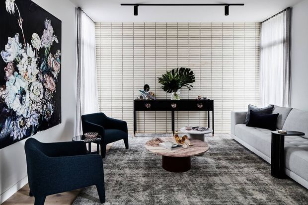 A refined contemporary living room with floor-to-ceiling sheer curtains, a white sectional sofa, two deep charcoal lounge chairs, a round marble coffee table, and a black console against a white horizontally lined feature wall. A large floral artwork provides a dramatic accent, while a textured grey rug and sculptural greenery soften the monochromatic scheme. Track lighting runs across the ceiling.