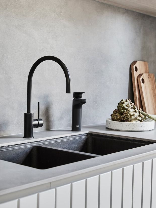 Close-up of a contemporary kitchen sink area featuring a matte black gooseneck tap and matching soap dispenser, a deep double-bowl black sink, pale grey benchtops, white vertical-groove cabinetry, and a textured grey wall. Wooden chopping boards and a plate with an artichoke sit on the benchtop.