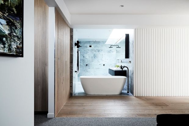 Bright contemporary bathroom viewed from an adjacent carpeted room, featuring a freestanding white bathtub, black tapware, a glass shower enclosure, pale textured wall tiles, timber flooring, vertical white wall panelling, and timber-lined cabinetry or wall surfaces. A partial black sofa or chair is visible in the foreground.