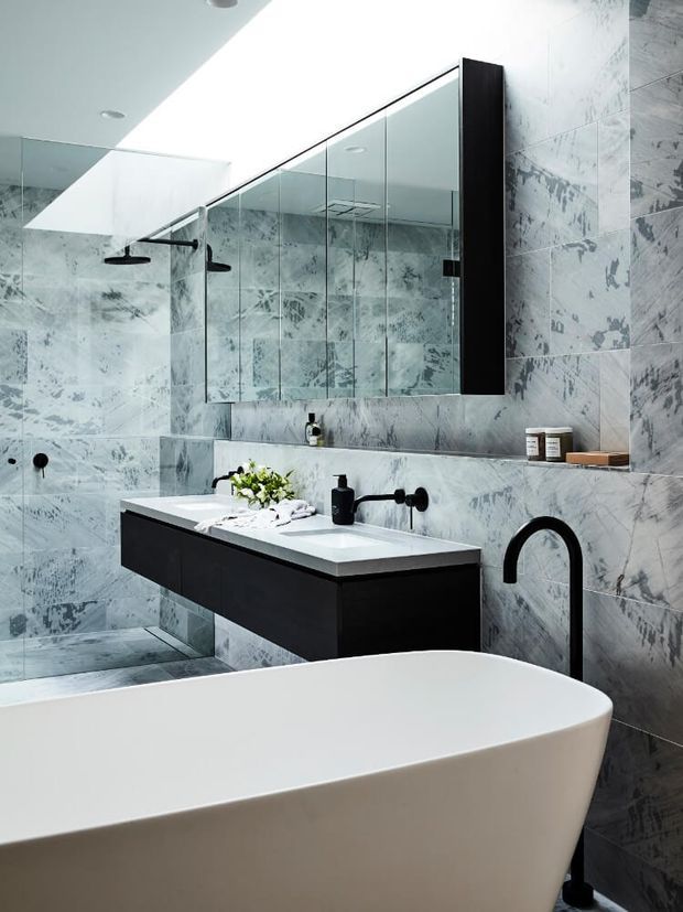 A sophisticated contemporary bathroom featuring a freestanding white bathtub in the foreground, a floating dark vanity with an integrated white basin, wall-mounted black tapware, a large frameless mirrored cabinet, and a glass-enclosed shower. Grey-white veined stone-look wall and floor tiles create a continuous marble-like finish, complemented by black fittings and joinery.