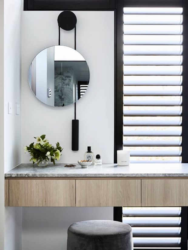 A refined bathroom vanity area with a floating light-wood cabinet, pale stone benchtop, large round mirror, black linear pendant detail, and black-framed horizontal louvred window. A dark upholstered stool and small decorative items sit below the vanity.