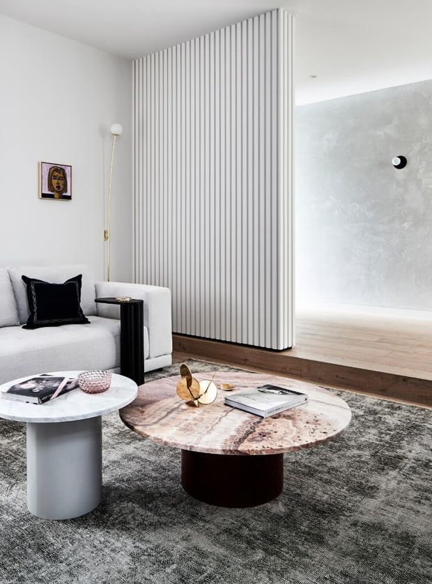 A refined contemporary living room with a white sofa, black accent cushion, two sculptural coffee tables, and a large textured rug. A dramatic floor-to-ceiling vertical slatted partition separates the seating area from an adjoining pale textured-wall space. The palette is predominantly white, grey, light timber and dark accents, with curated artwork and metallic decor.