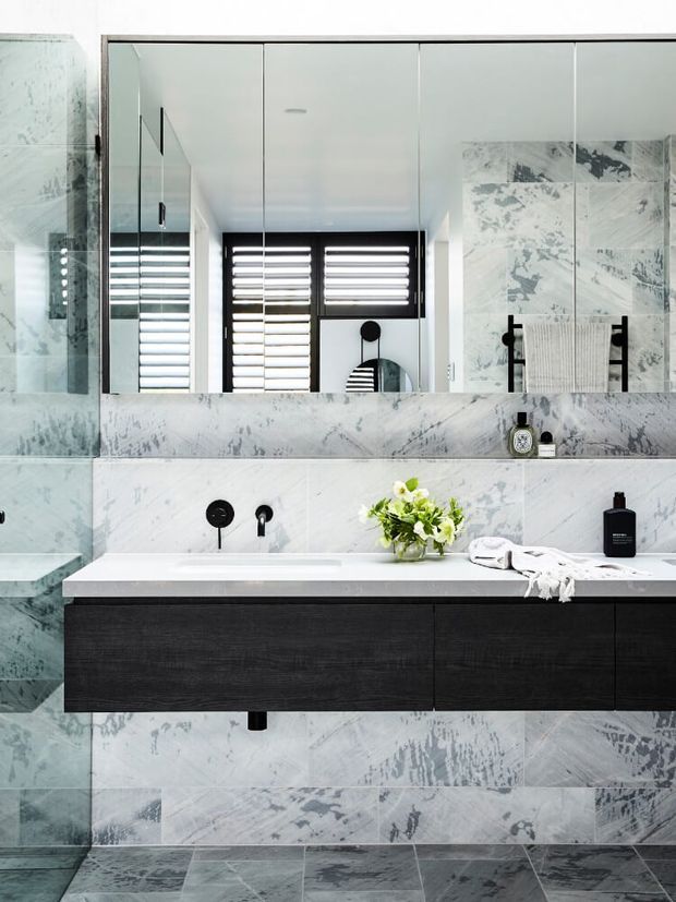 Contemporary bathroom featuring a floating dark timber vanity with an integrated white countertop, wall-to-wall pale grey veined stone or marble-look tiles, a large mirrored cabinet, black fixtures, and a frameless glass shower enclosure. Plantation shutters and a small arrangement of greenery add detail to the clean, monochromatic scheme.