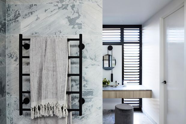 Contemporary bathroom featuring a black heated towel rail mounted against large pale marble-look wall tiles. The adjacent vanity has a timber-fronted drawer, white countertop, round mirror, black wall-mounted fittings, a grey upholstered stool, and tall black-framed louvred windows providing filtered light.